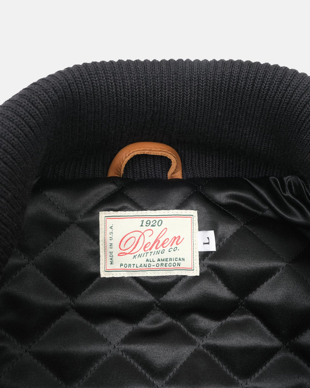 Close-up of the inside collar of a black quilted jacket or bag, showing a ribbed black knit collar, a tan leather handle loop, and a sewn-in label reading 'Dehen Knitting Co.'.
