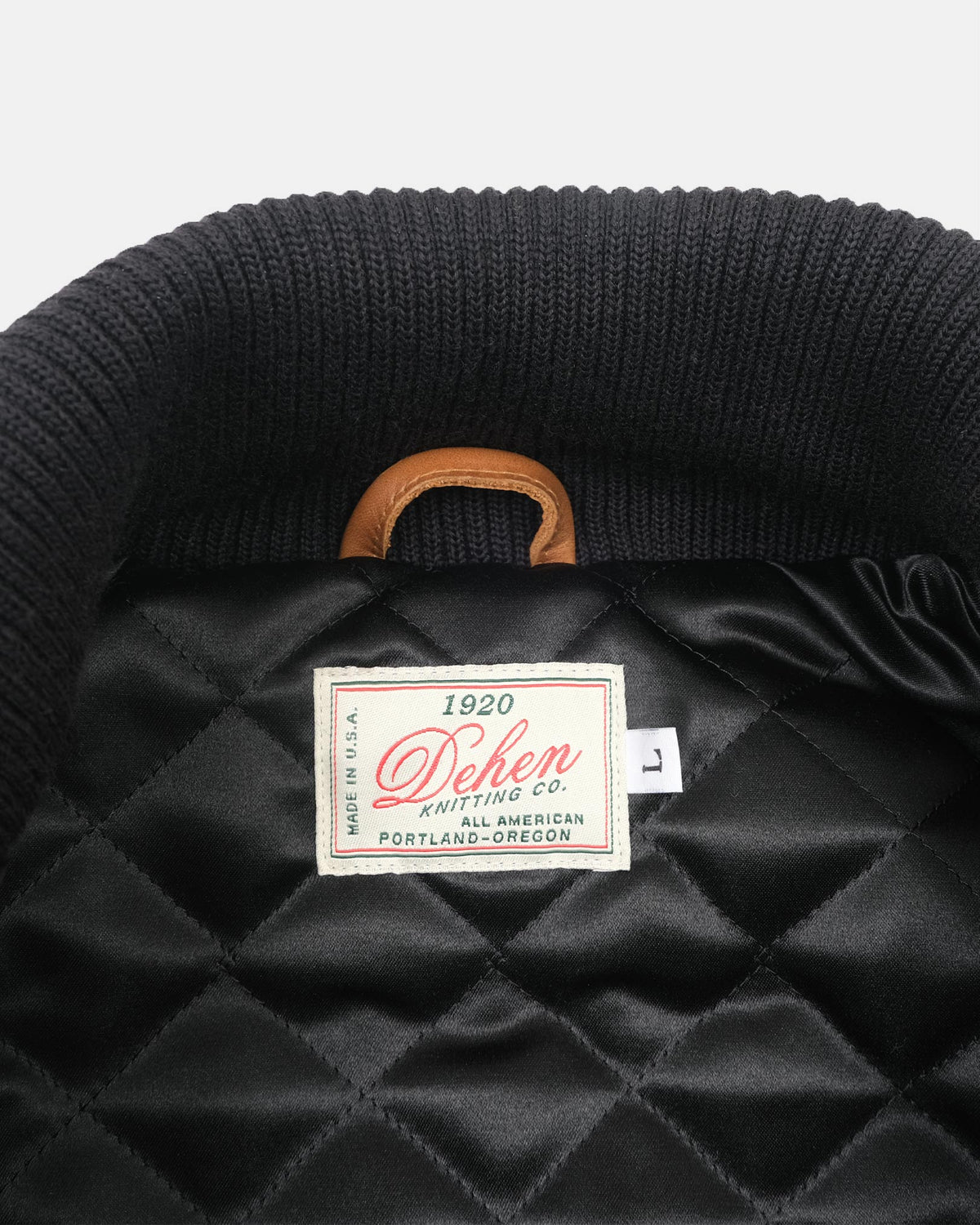 Close-up of the inside collar of a black quilted jacket or bag, showing a ribbed black knit collar, a tan leather handle loop, and a sewn-in label reading 'Dehen Knitting Co.'.