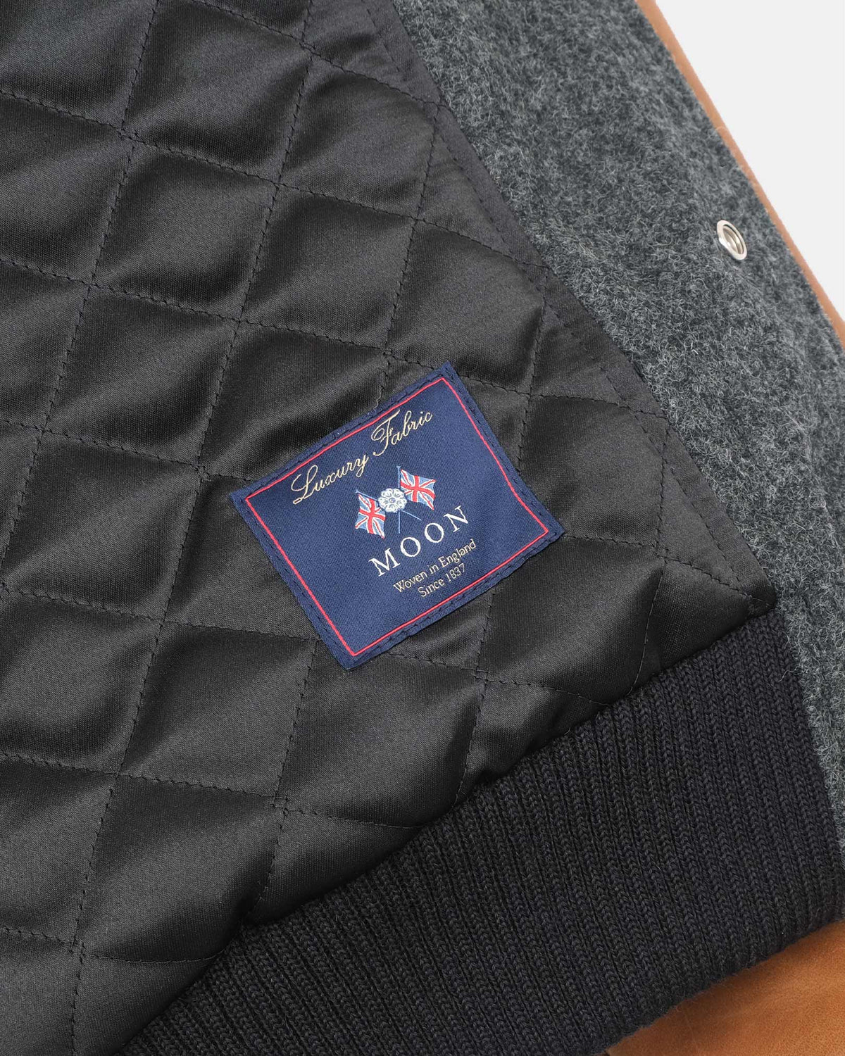 Close-up of a quilted dark gray jacket interior with a visible sewn label reading Luxury Fabrics MOON, alongside a ribbed dark knit collar and textured gray outer fabric.