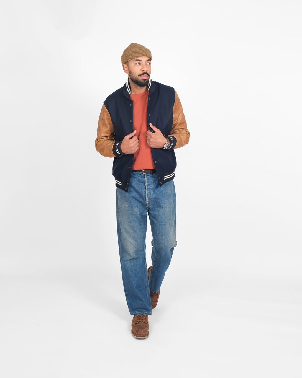 Man wearing a navy varsity jacket with brown sleeves, red shirt, blue jeans, and brown shoes on a white background