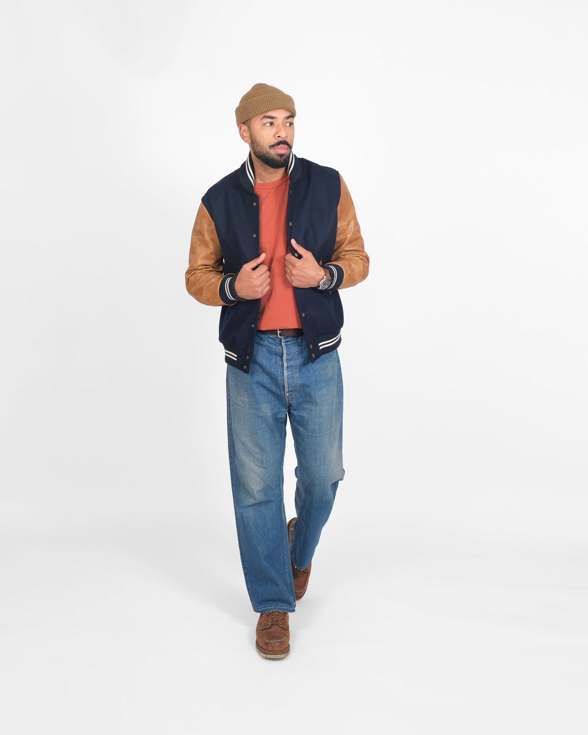Man wearing a navy varsity jacket with brown sleeves, red shirt, blue jeans, and brown shoes on a white background