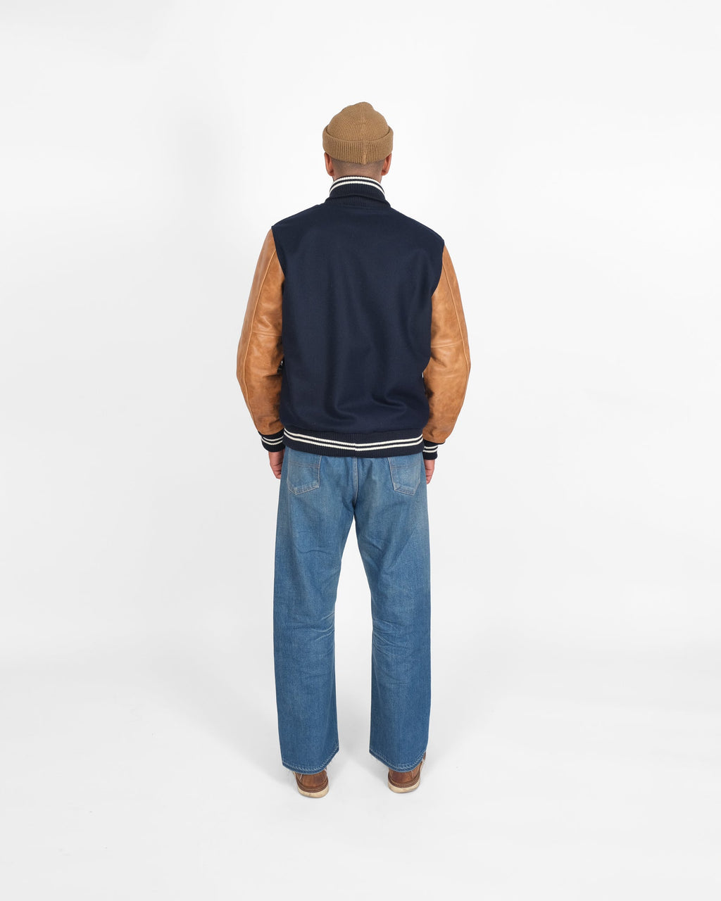 Person wearing a navy varsity jacket with brown sleeves and blue jeans on a white background