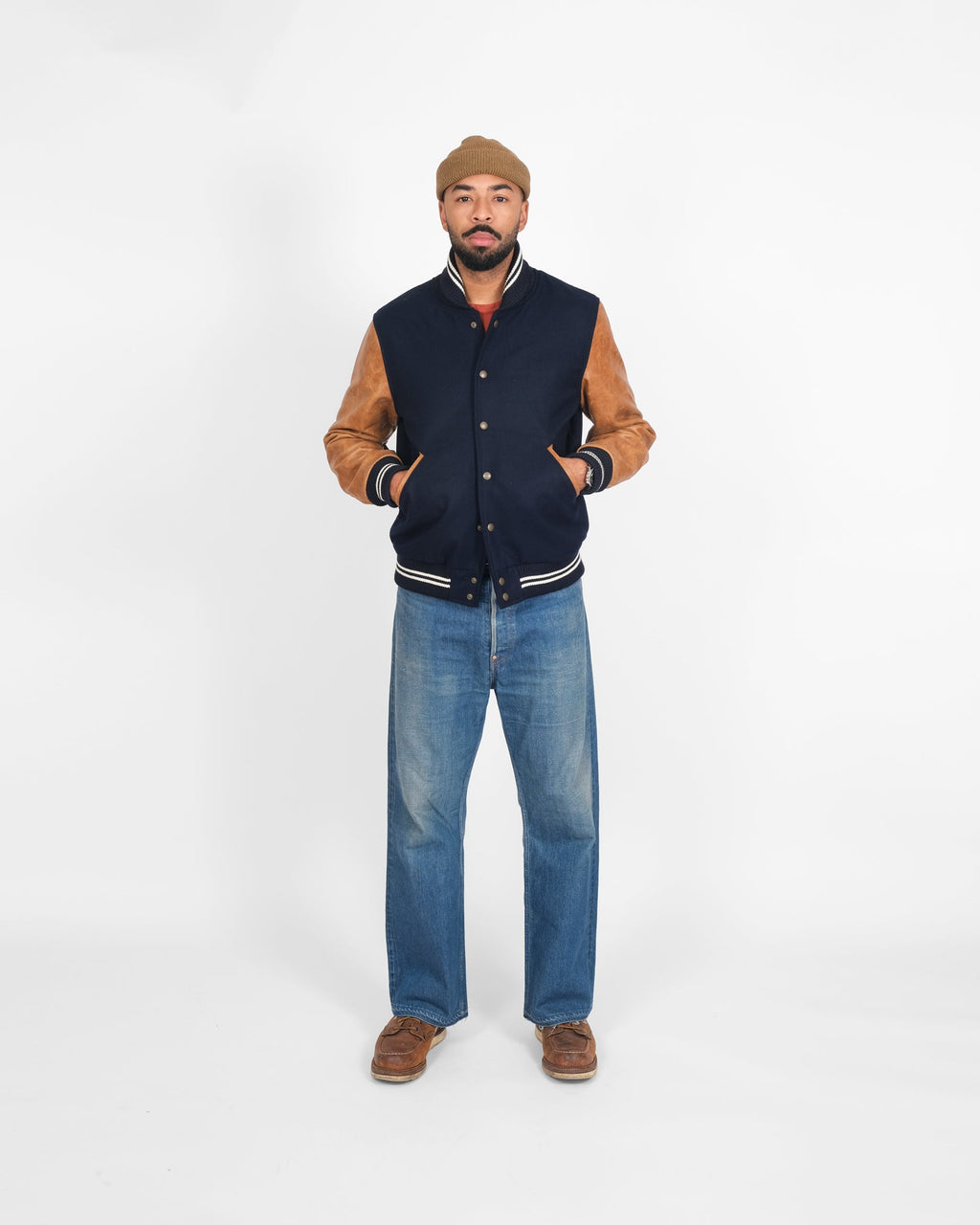 Man wearing a navy and brown varsity jacket with blue jeans on a white background