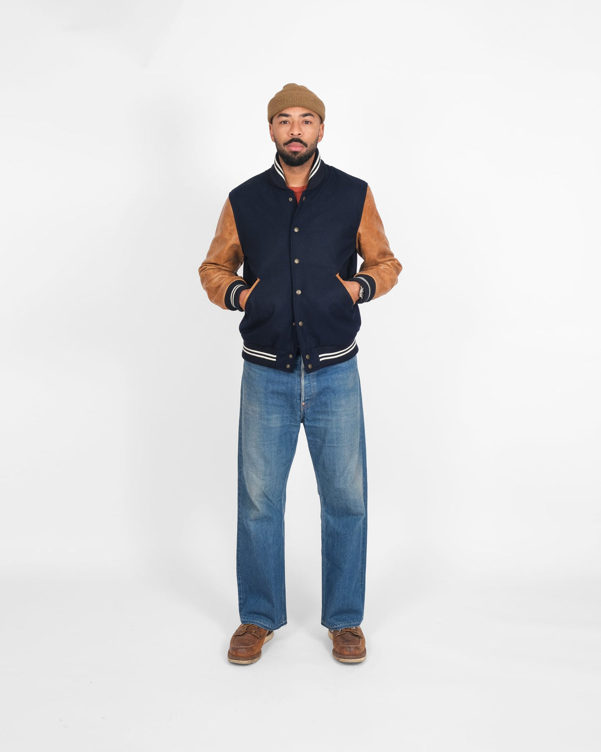 Man wearing a navy and brown varsity jacket with blue jeans on a white background