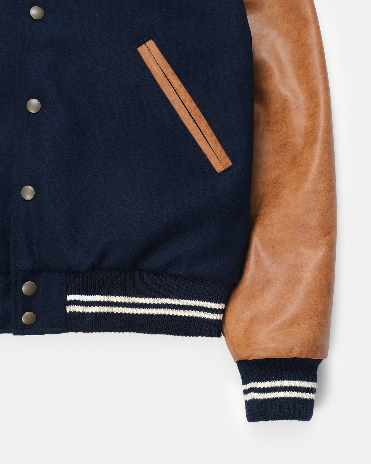 Close-up of a varsity-style jacket showing a navy blue body, tan leather sleeves, a diagonal tan welt pocket, and navy ribbed cuffs with white stripes on a light gray background.