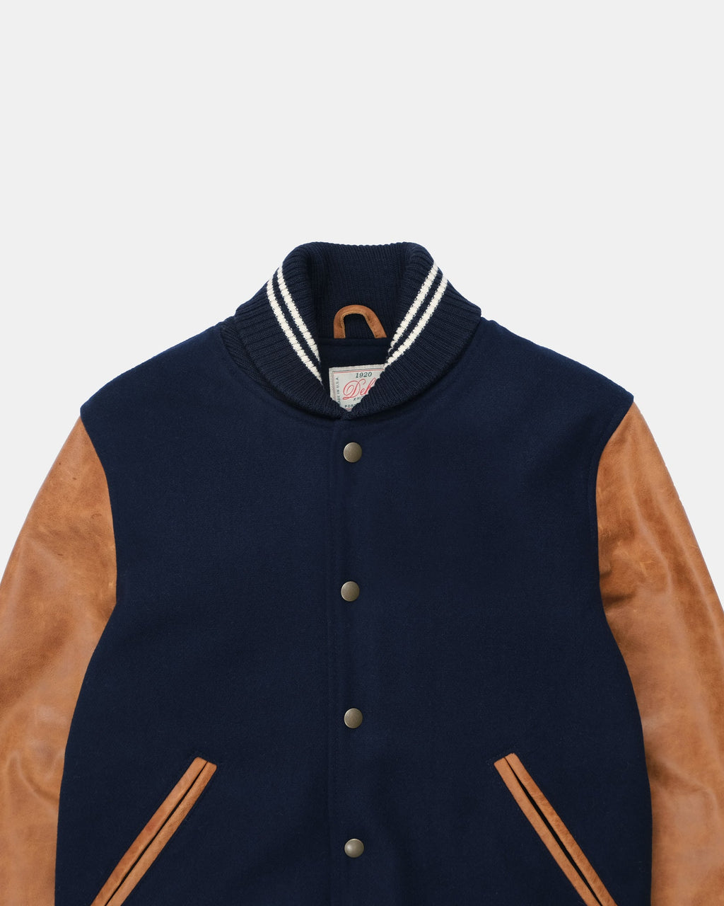A close-up of a varsity-style jacket with a navy blue body, tan suede sleeves, and a ribbed collar with white and navy stripes.