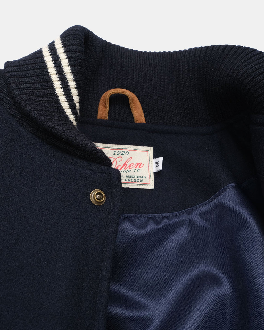 Close-up of a varsity-style jacket collar and upper chest showing a navy knit ribbed collar with white stripes, a tan leather handle loop, and a navy satin-lined front panel.