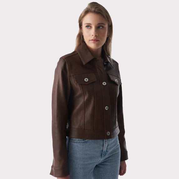 A woman wearing a dark brown cropped leather jacket with silver snap buttons and blue jeans, photographed against a light gray studio background.