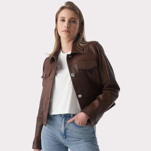 A woman wearing a brown cropped leather jacket over a white t-shirt and blue jeans, photographed against a solid light gray background.