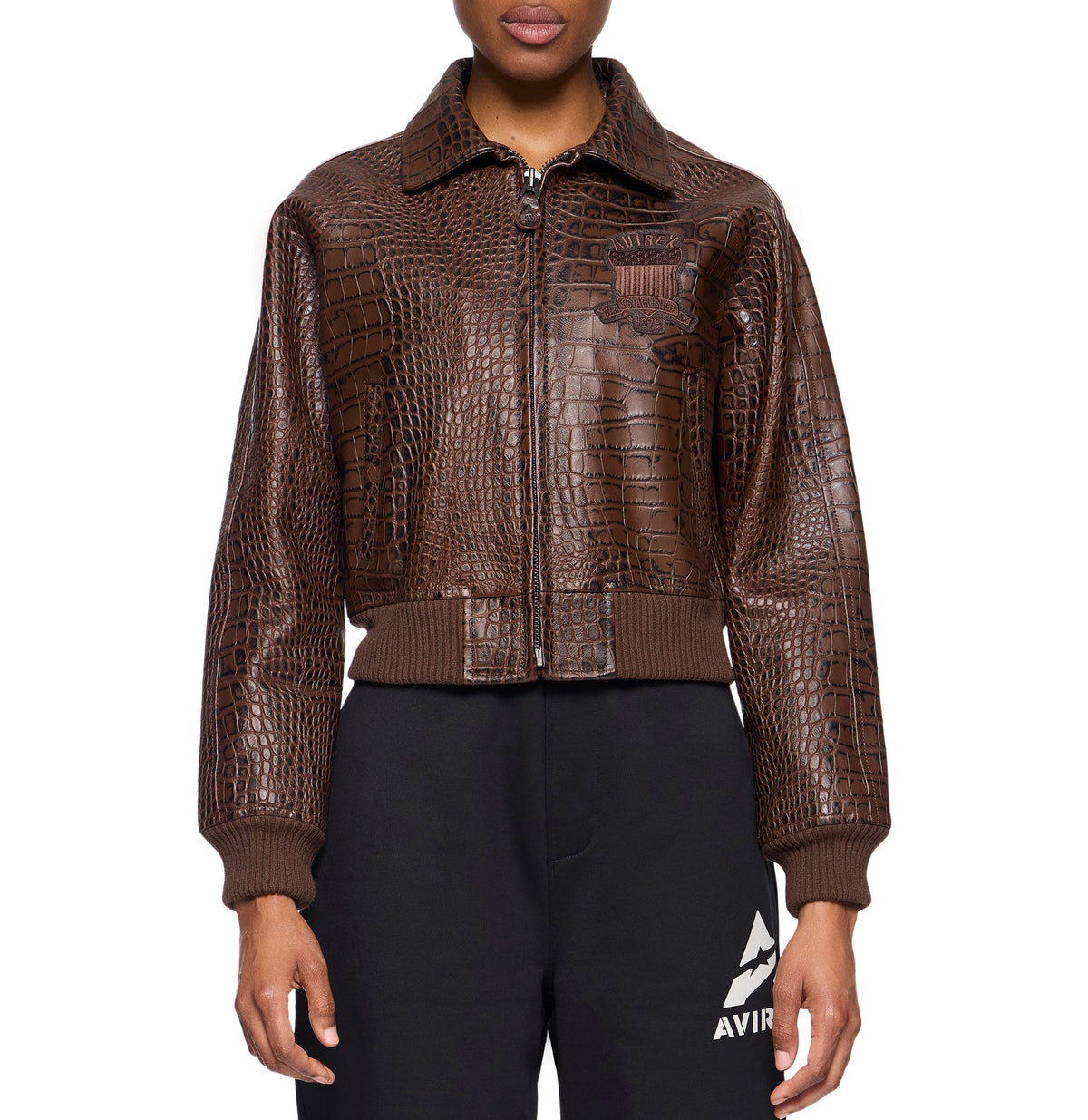 A model wearing a brown crocodile-embossed leather bomber jacket with ribbed knit collar, cuffs, and hem, paired with black sweatpants featuring a white logo.