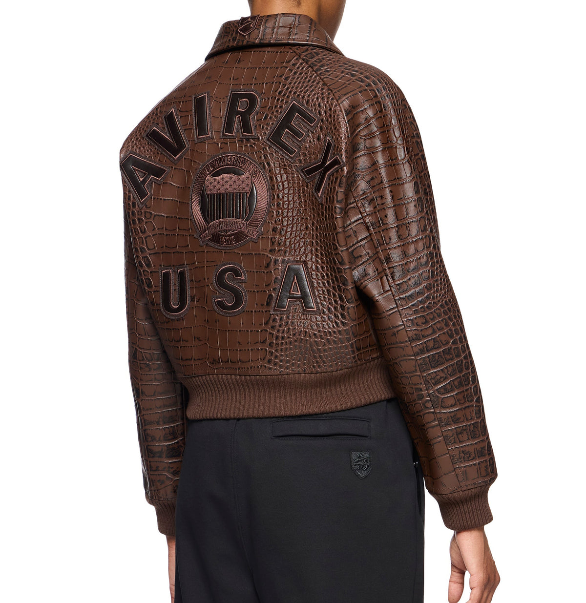 A model shown from the back wearing a brown crocodile-embossed leather bomber jacket with 'AVIREX USA' and a crest patch on the back, paired with black trousers.