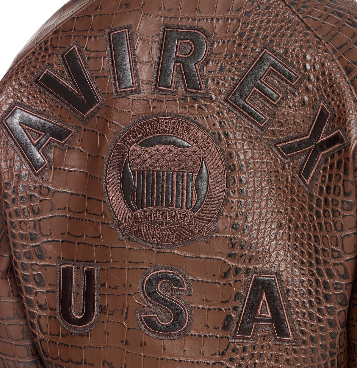 Close-up of a brown textured leather jacket featuring embossed crocodile skin pattern, large raised lettering reading 'AVIREX' and 'USA', and a central crest patch with an eagle motif and the words 'CROW AMERICAN EST. 1902'.