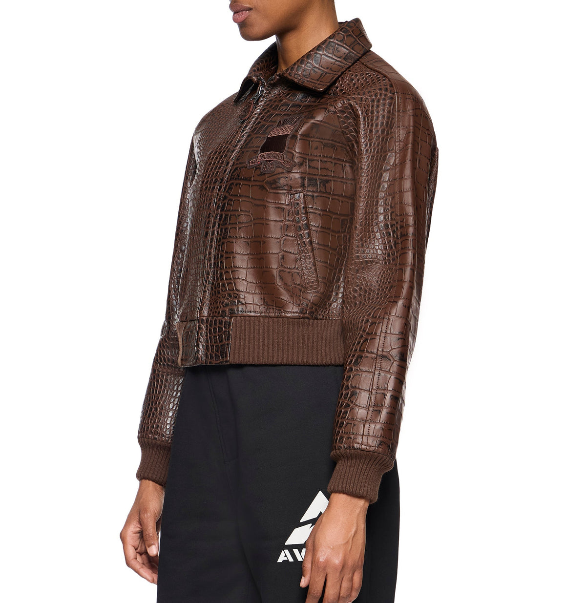 A model wearing a brown textured leather jacket with a crocodile pattern, ribbed knit collar, cuffs, and hem, paired with black sweatpants against a white background.