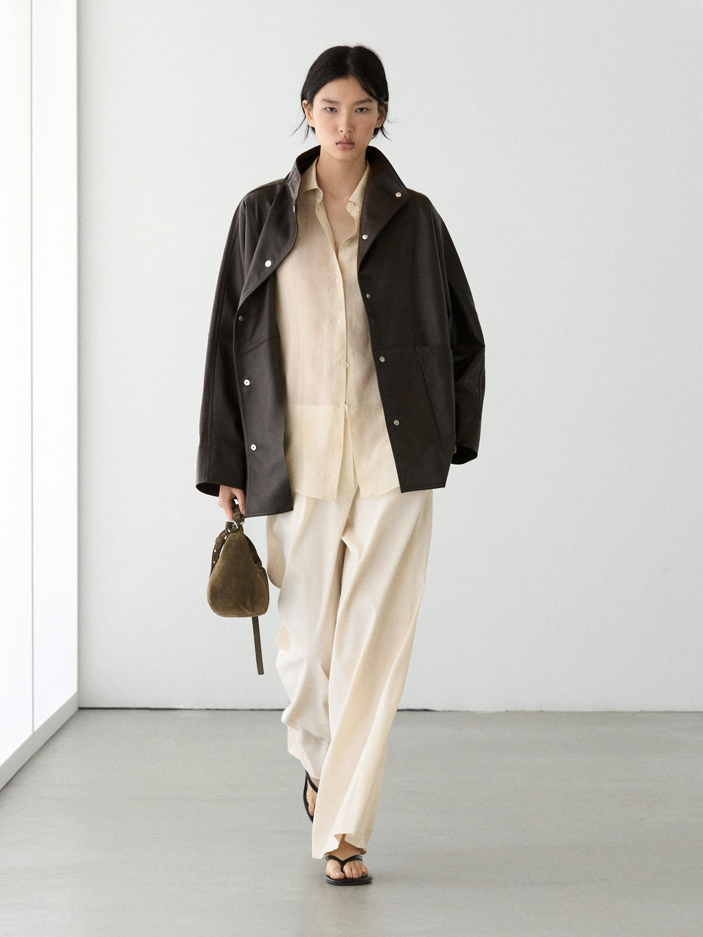 A model walks in a minimalist studio wearing a black leather jacket over a cream shirt and wide-leg cream trousers, holding a small brown handbag.