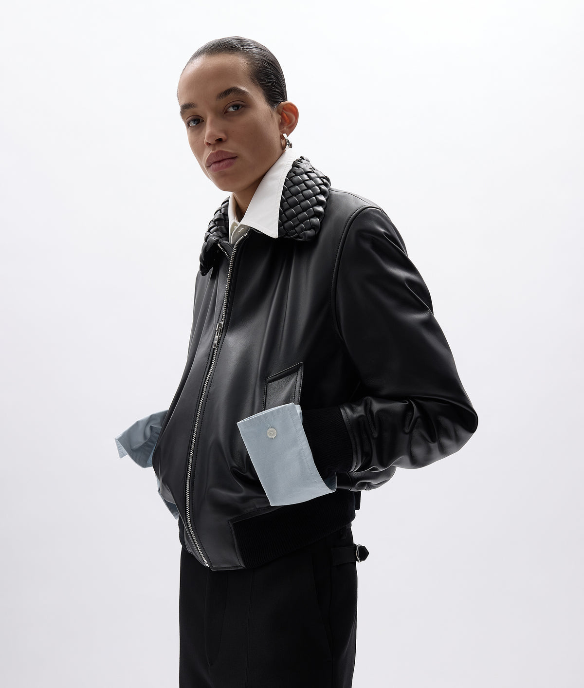 A model wearing a black leather bomber jacket with a chunky black knit collar, a white collared shirt underneath, and light blue fabric cuffs peeking out from the sleeves, set against a plain light gray studio background.