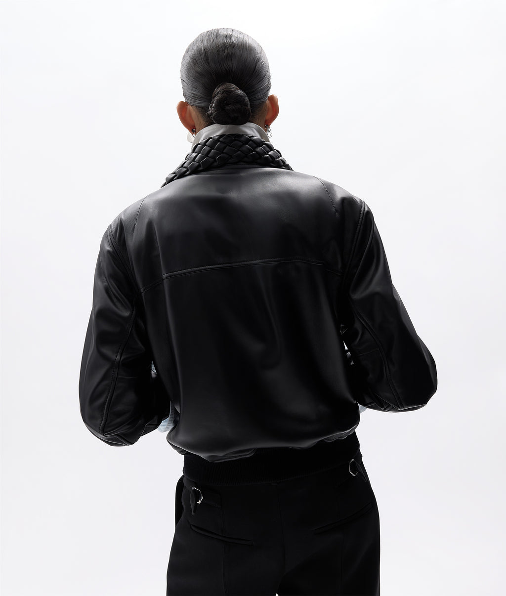 A person shown from the back wearing a black faux-leather jacket with a chunky black knit collar, paired with black trousers, against a solid white background.