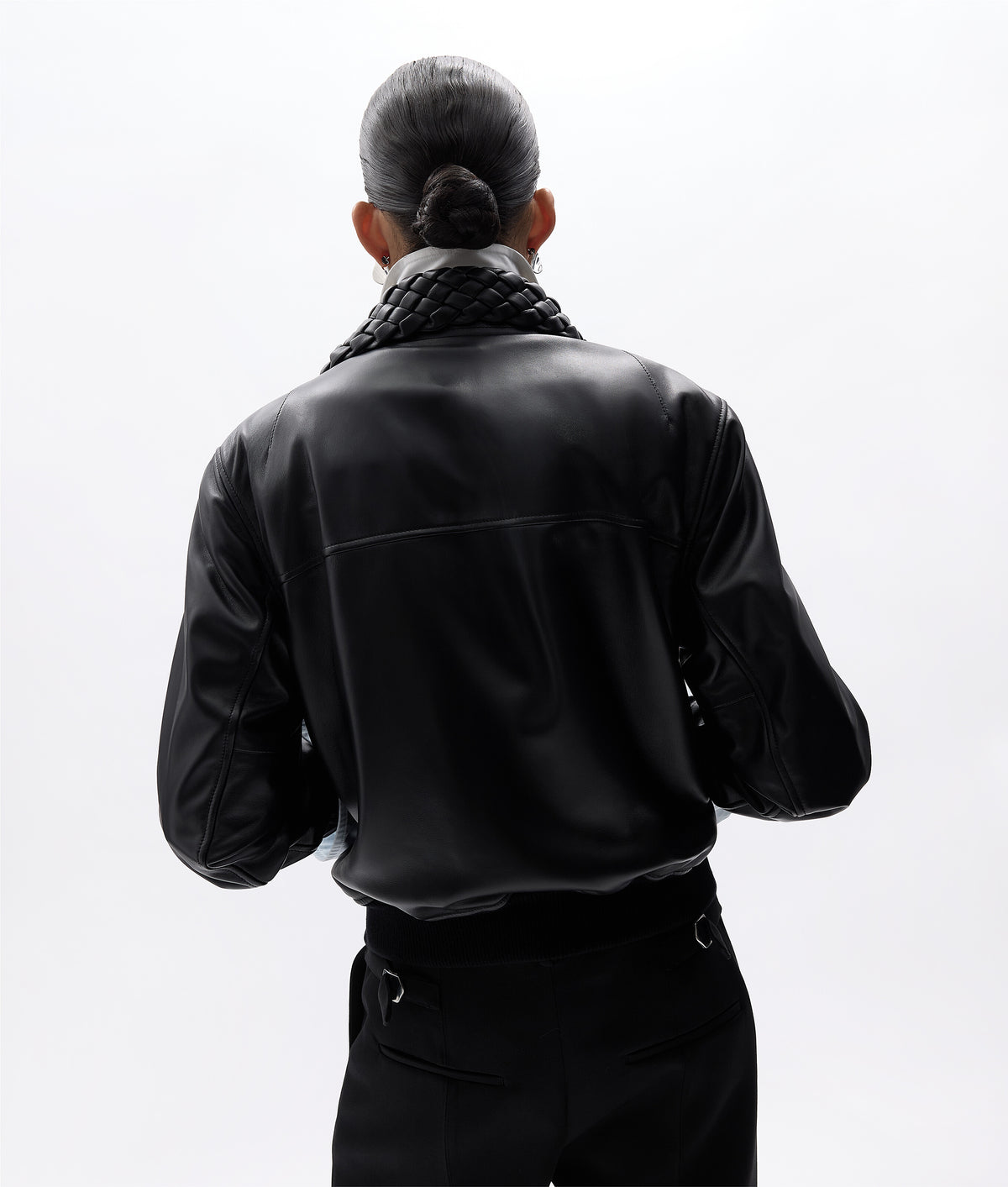 A person shown from the back wearing a black faux-leather jacket with a chunky black knit collar, paired with black trousers, against a solid white background.