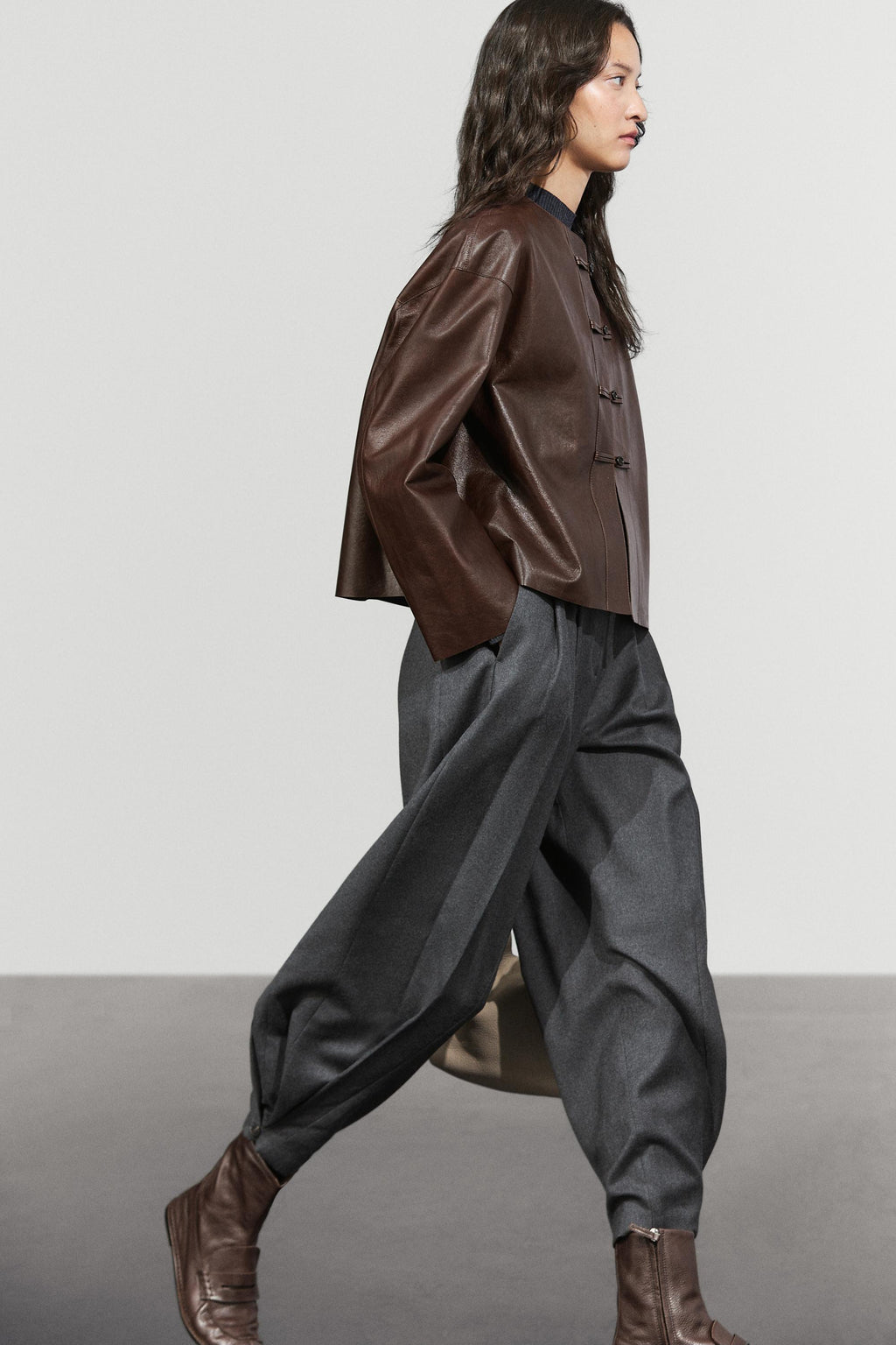 A model walks in profile wearing a dark brown leather jacket with a mandarin collar and button closure, paired with loose dark gray baggy trousers and brown leather boots, set against a minimalist studio background.