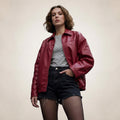 A woman wearing a red leather jacket over a gray and white striped t-shirt, paired with black denim shorts and sheer black tights, standing against a plain beige background.