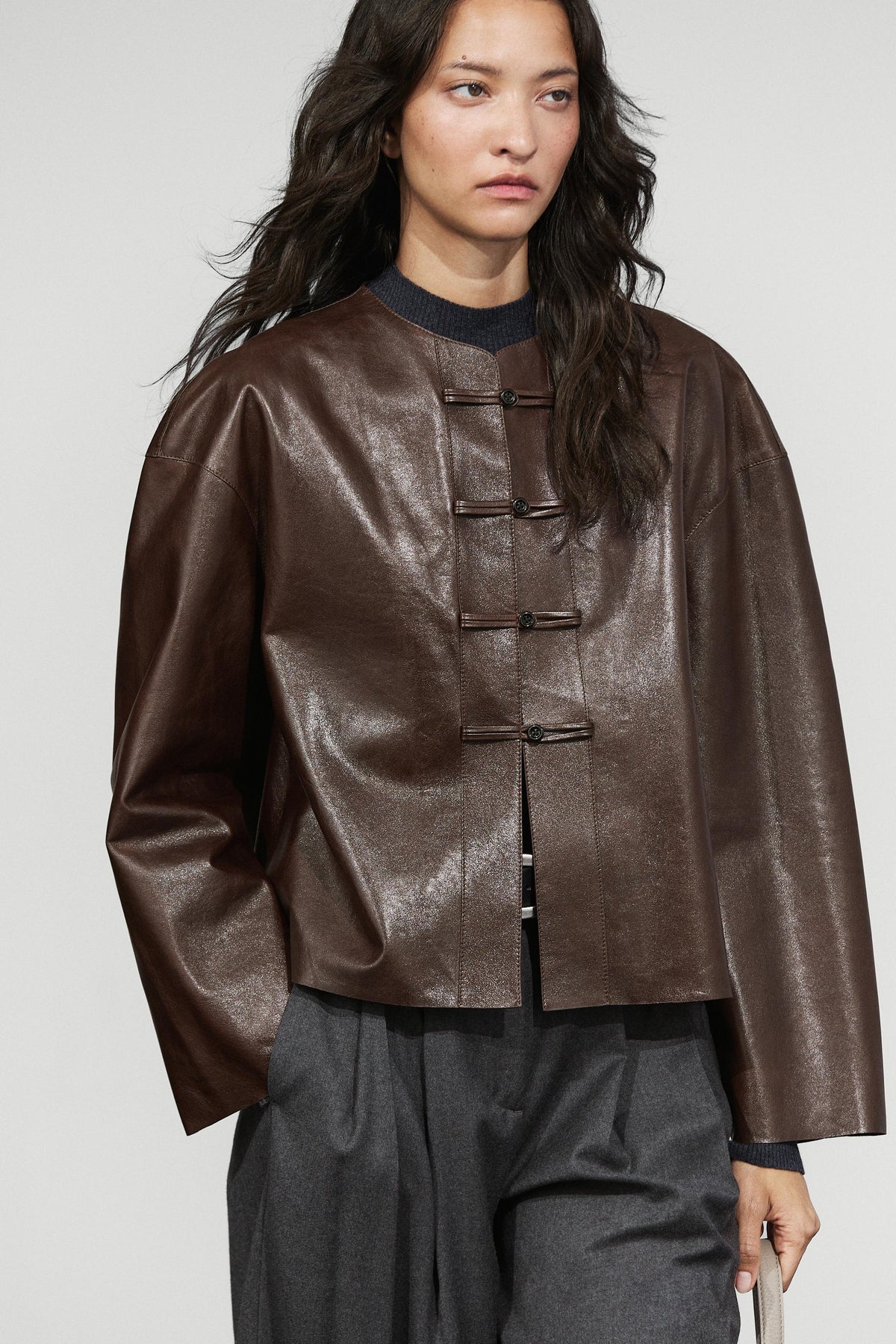 A woman wearing a cropped, long-sleeve brown faux-leather jacket with traditional Chinese-style frog closures, paired with dark gray trousers, against a light gray studio background.