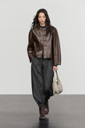 A model walks down a runway wearing a dark brown cropped leather jacket with toggle closures, loose dark gray baggy pants, brown boots, and holding a light beige tote bag.