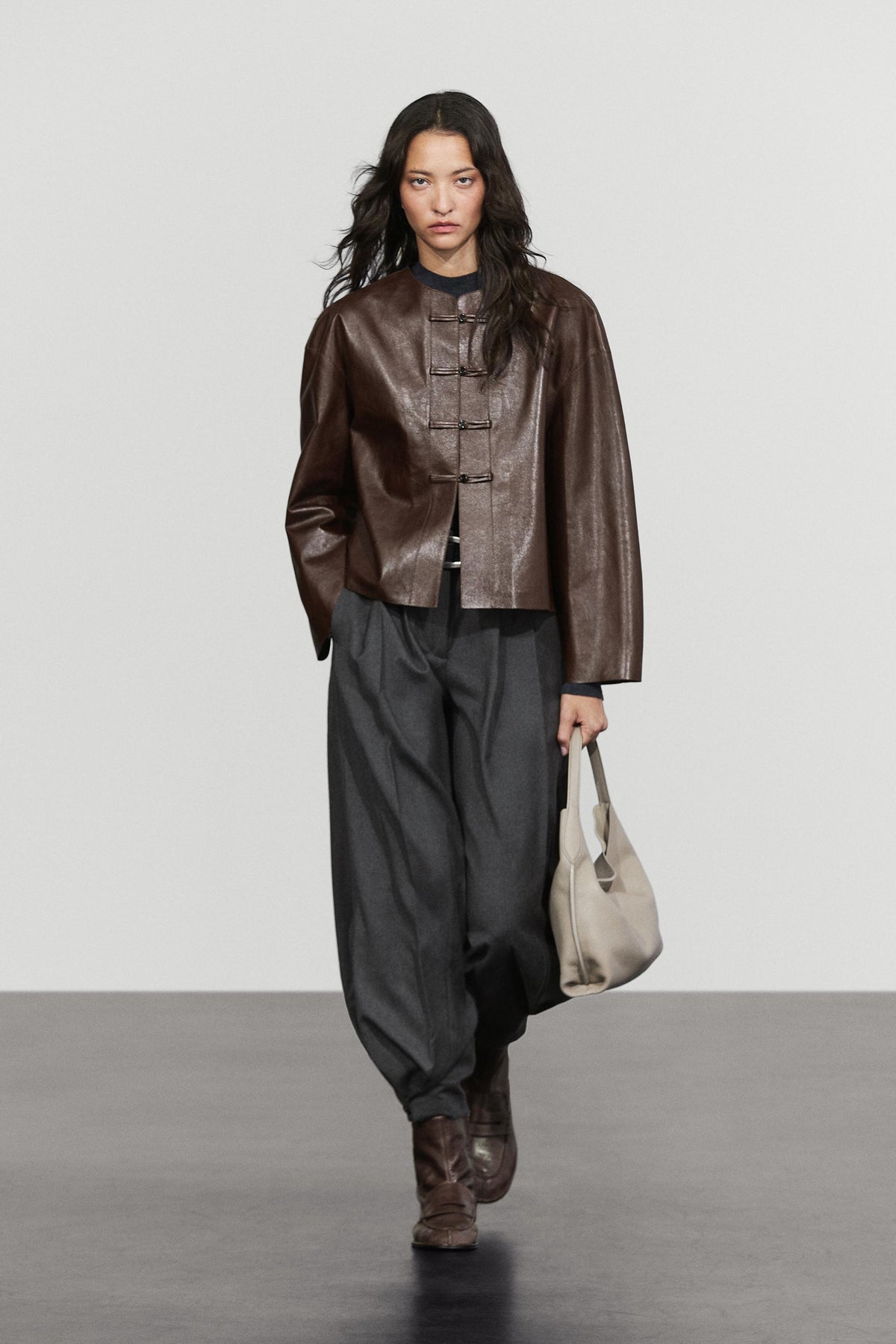 A model walks down a runway wearing a dark brown cropped leather jacket with toggle closures, loose dark gray baggy pants, brown boots, and holding a light beige tote bag.