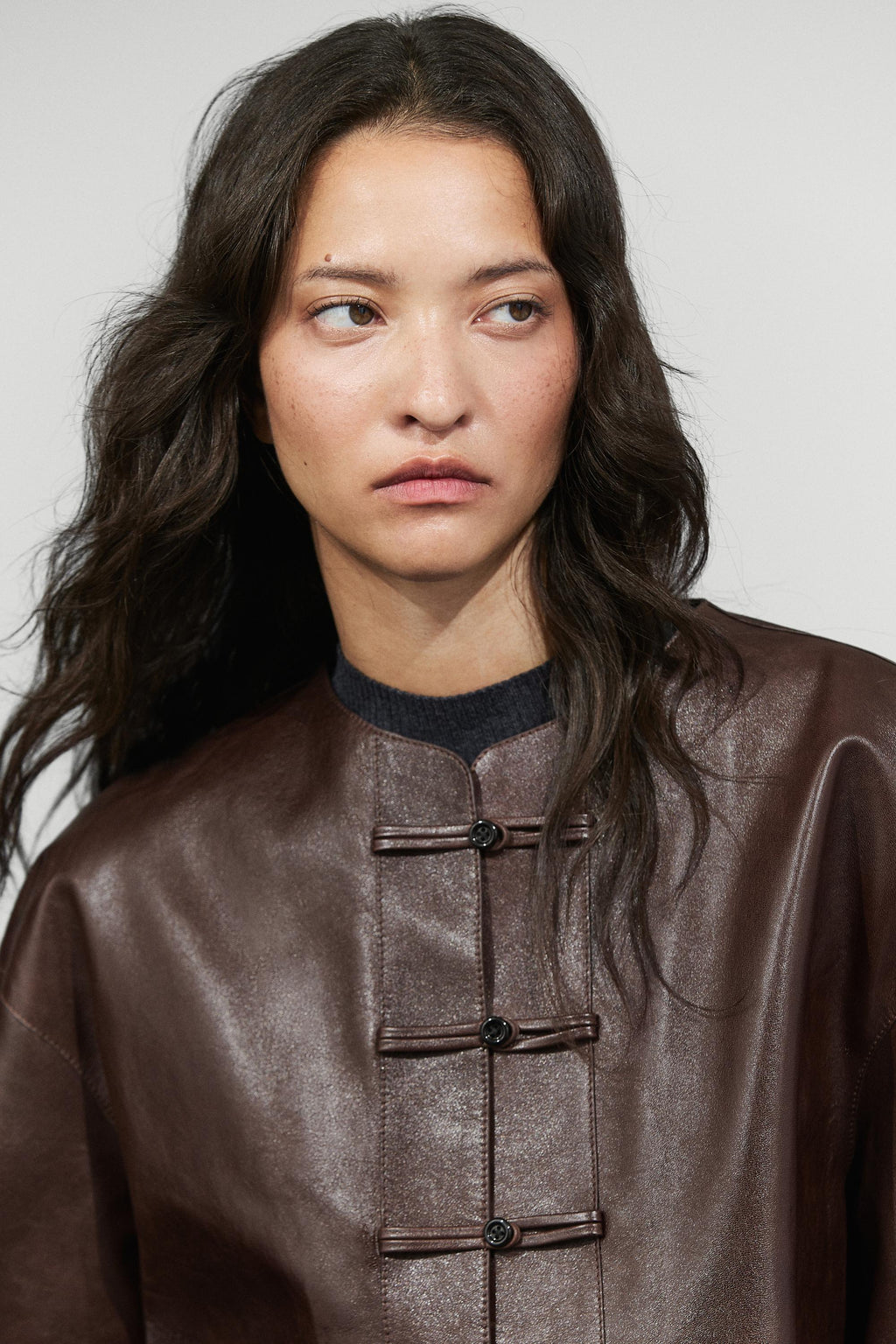 A close-up portrait of a model wearing a dark brown leather jacket with traditional Chinese-style frog closures and a dark crew neck.