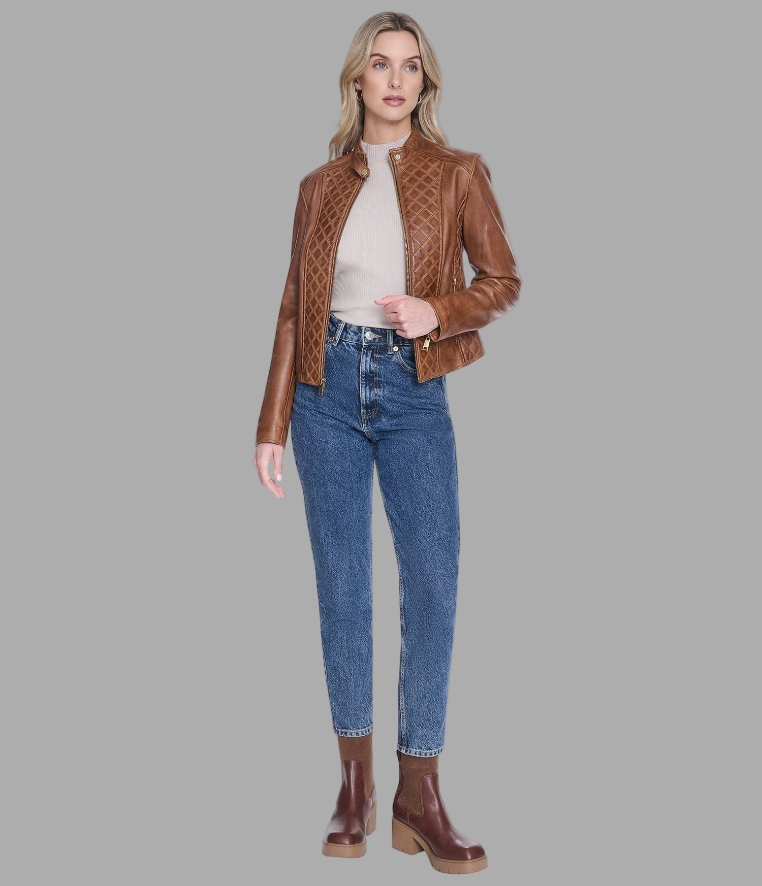 A woman modeling a brown quilted leather jacket, styled with a cream top, blue jeans, and brown leather boots, against a solid gray background.
