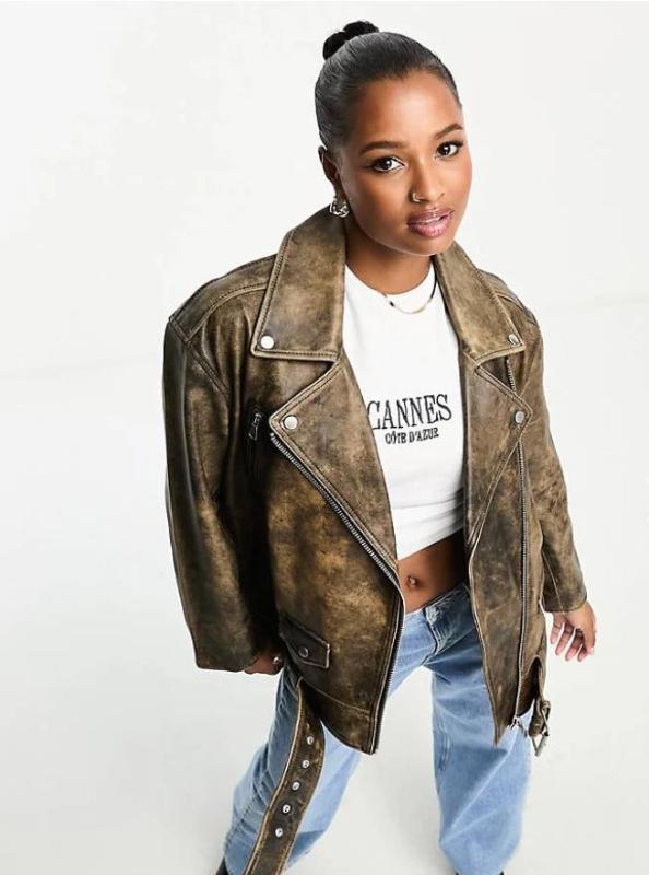 A woman wearing a distressed brown faux-leather moto jacket over a white cropped graphic t-shirt and light blue jeans, photographed against a plain white studio background.
