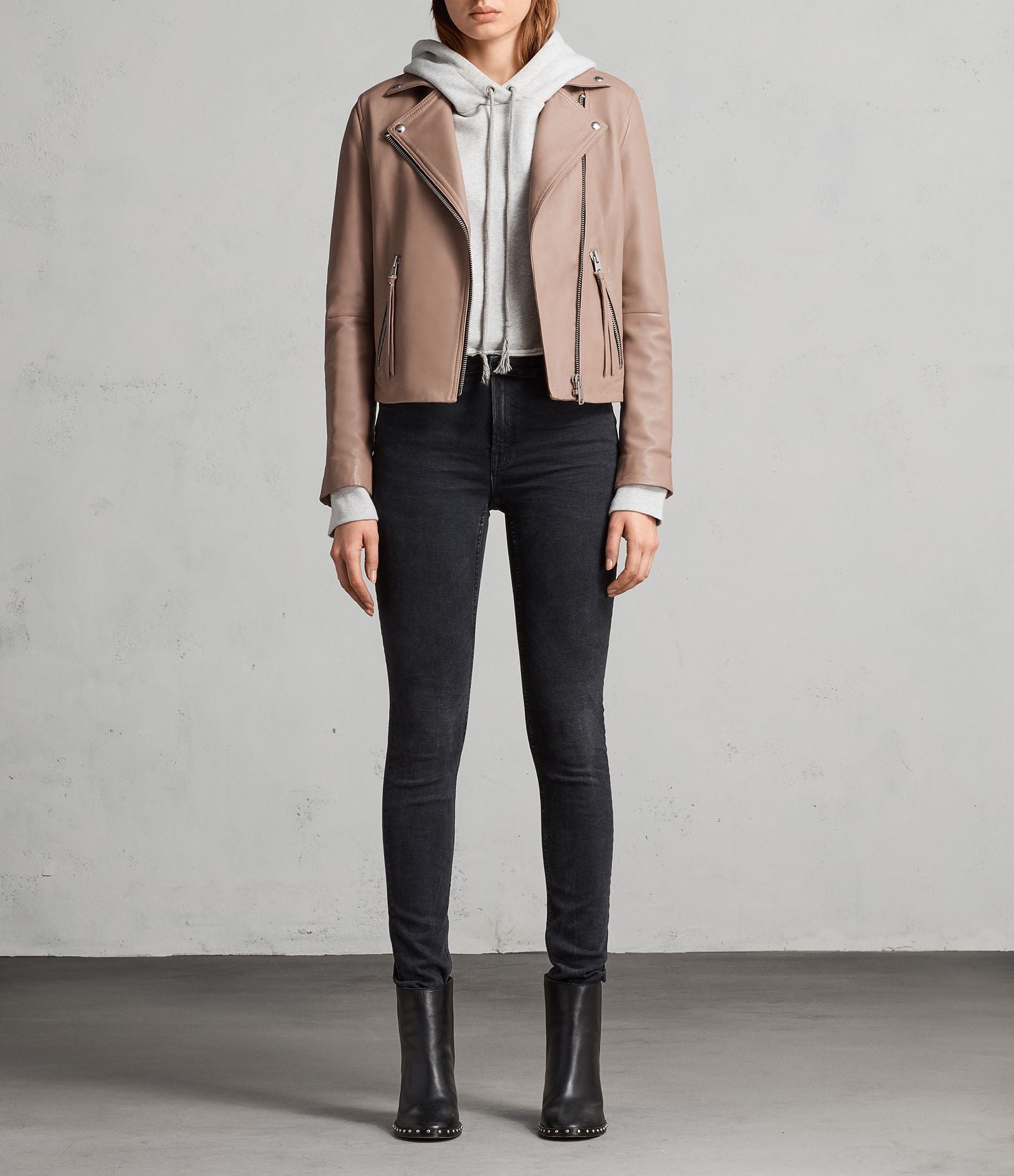 Person wearing a beige jacket, white hoodie, black jeans, and black boots against a gray background