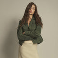 A woman wearing a cropped olive green suede moto jacket and a cream-colored skirt, posing with her arms crossed against a plain, light gray studio background.