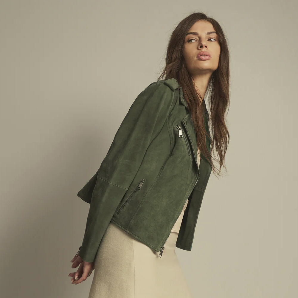 A woman wearing an olive green suede moto jacket with silver zippers, styled over a cream-colored top and skirt, photographed against a neutral beige studio background.