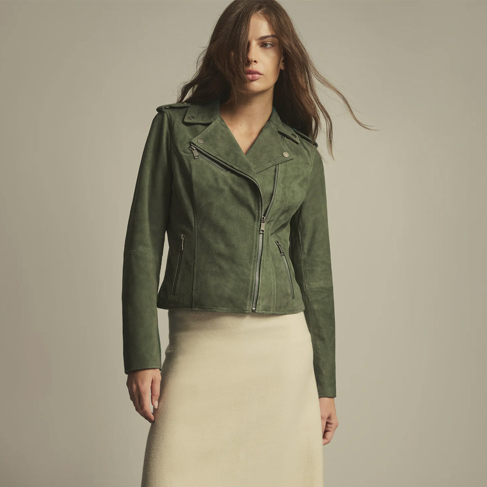 A woman modeling a cropped, olive green suede moto jacket with an asymmetrical zipper, paired with a cream-colored skirt, against a solid light gray studio background.