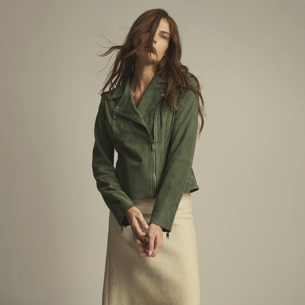 A woman wearing a muted olive green moto jacket and a cream-colored skirt, posing against a solid light gray studio background.