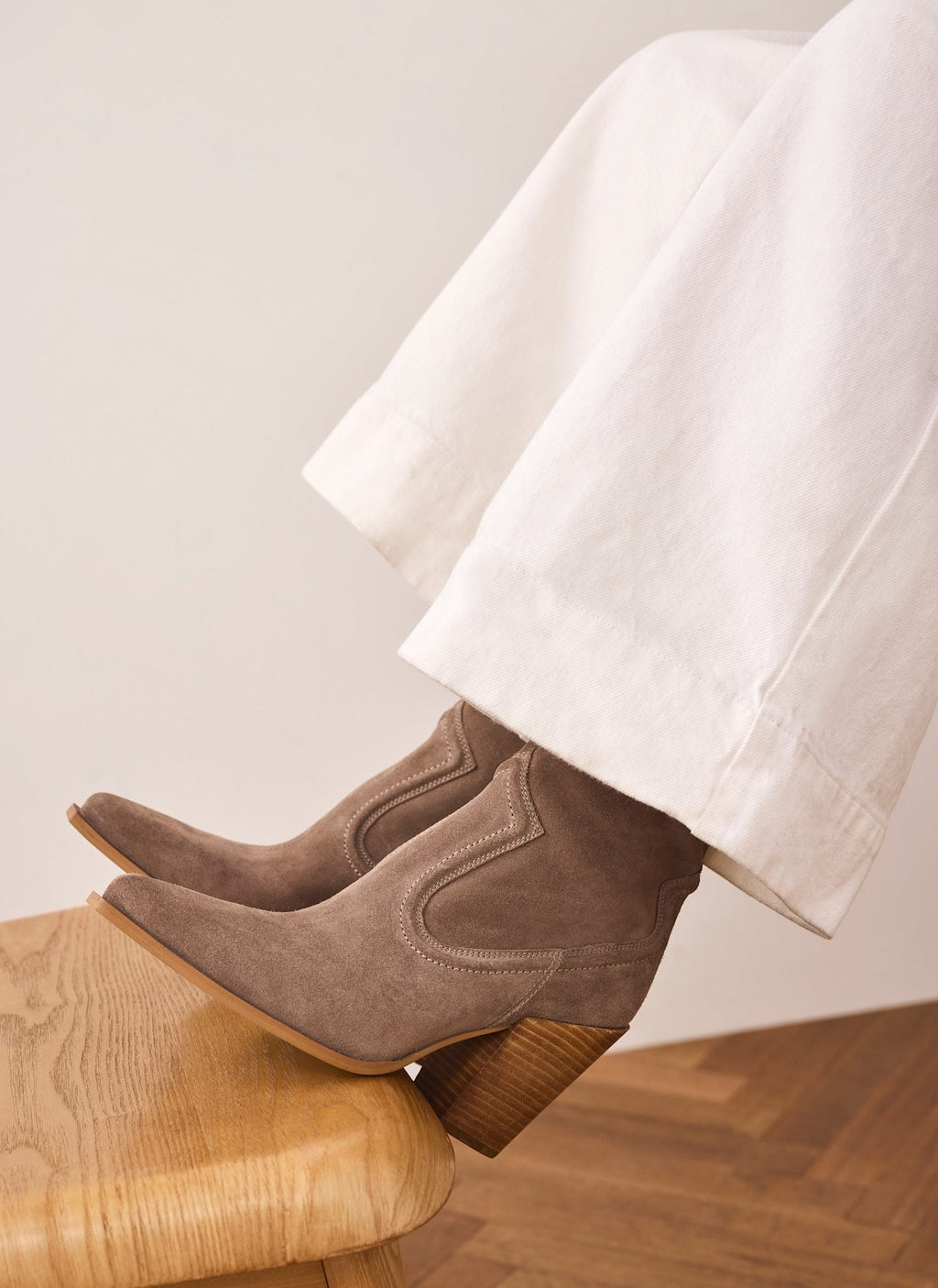 A pair of taupe-brown suede ankle boots with chunky stacked wood-look heels, shown on a model's feet resting on a wooden stool. The model is wearing wide-leg white trousers against a neutral beige wall and wood floor.