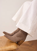 A pair of taupe-brown suede ankle boots with chunky stacked wood-look heels, shown on a model's feet resting on a wooden stool. The model is wearing wide-leg white trousers against a neutral beige wall and wood floor.