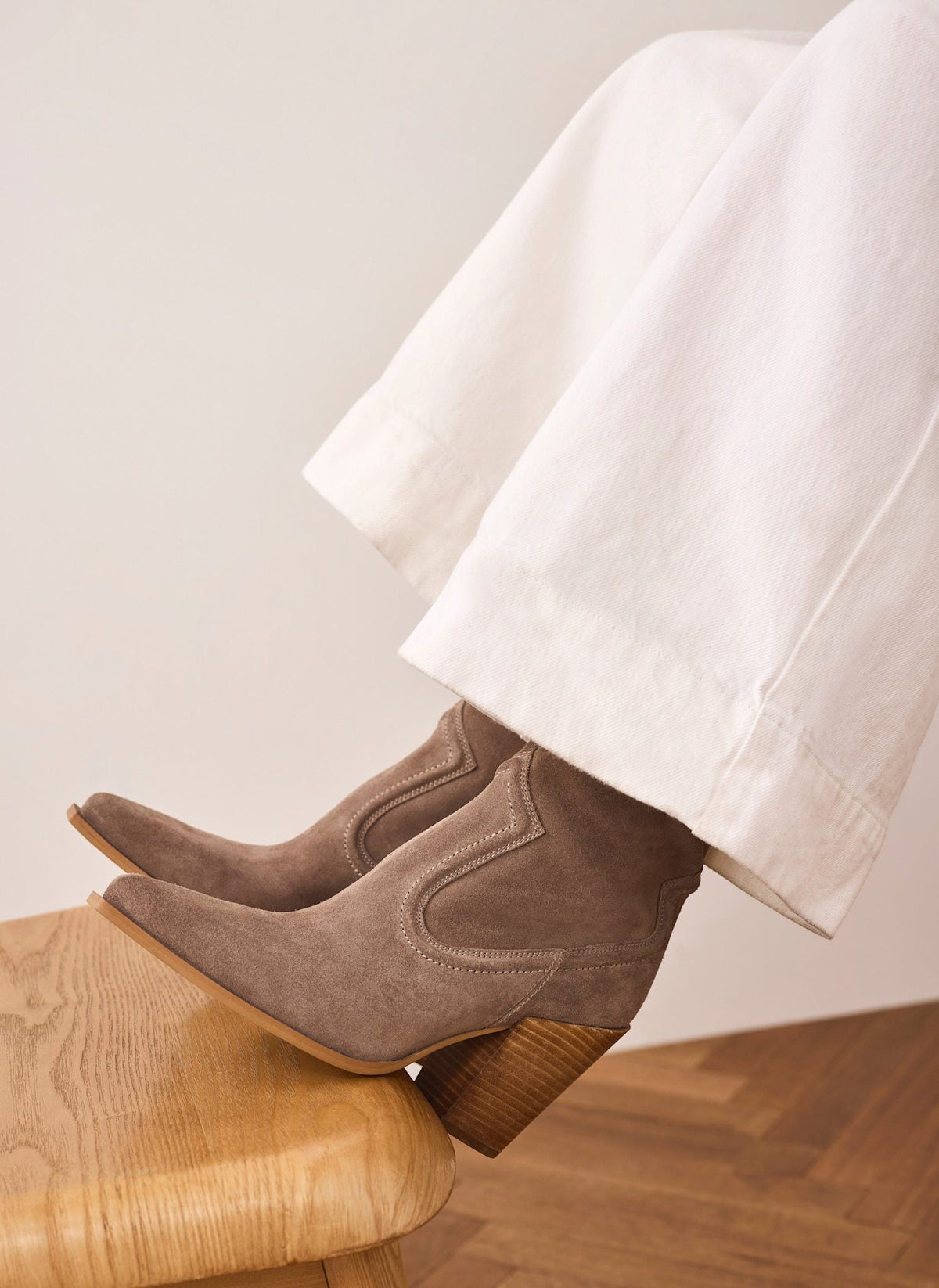 A pair of taupe-brown suede ankle boots with chunky stacked wood-look heels, shown on a model's feet resting on a wooden stool. The model is wearing wide-leg white trousers against a neutral beige wall and wood floor.
