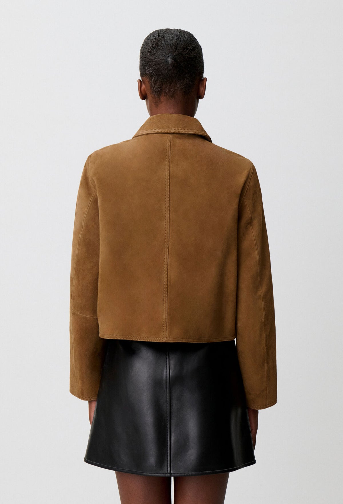 A model shown from the back wearing a cropped tan suede jacket and a black leather mini skirt against a light gray studio background.