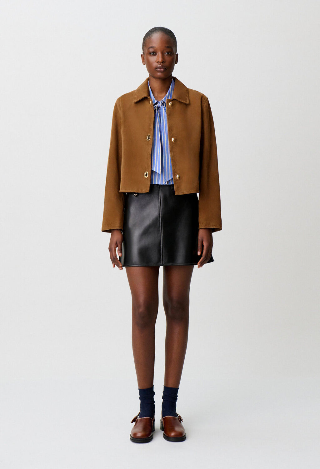 A model stands against a light gray studio background wearing a cropped brown suede jacket over a blue striped shirt, paired with a black leather mini skirt, dark socks, and brown leather loafers.
