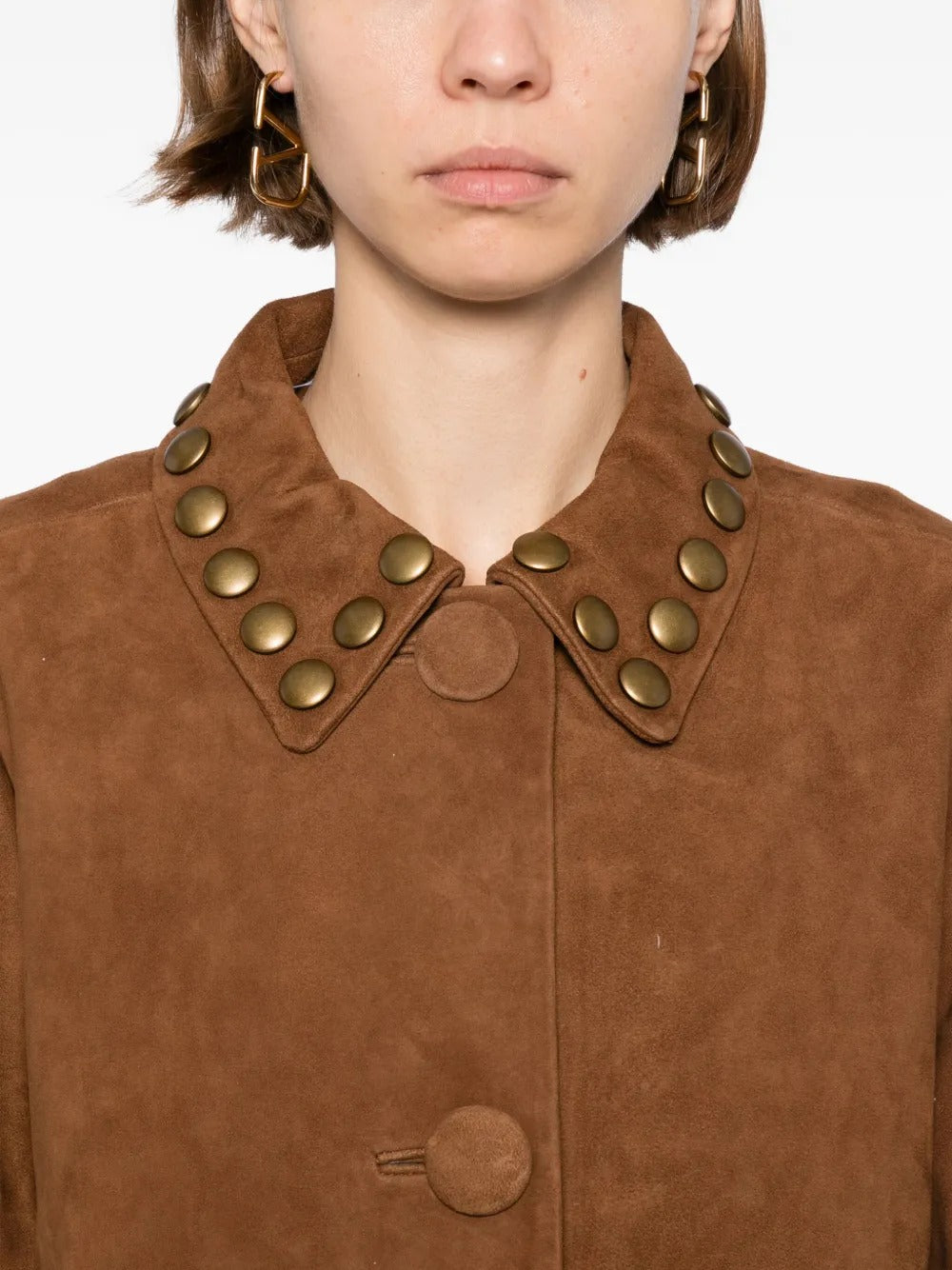 A close-up of a model wearing a brown suede jacket with a wide collar adorned with round metallic gold studs.