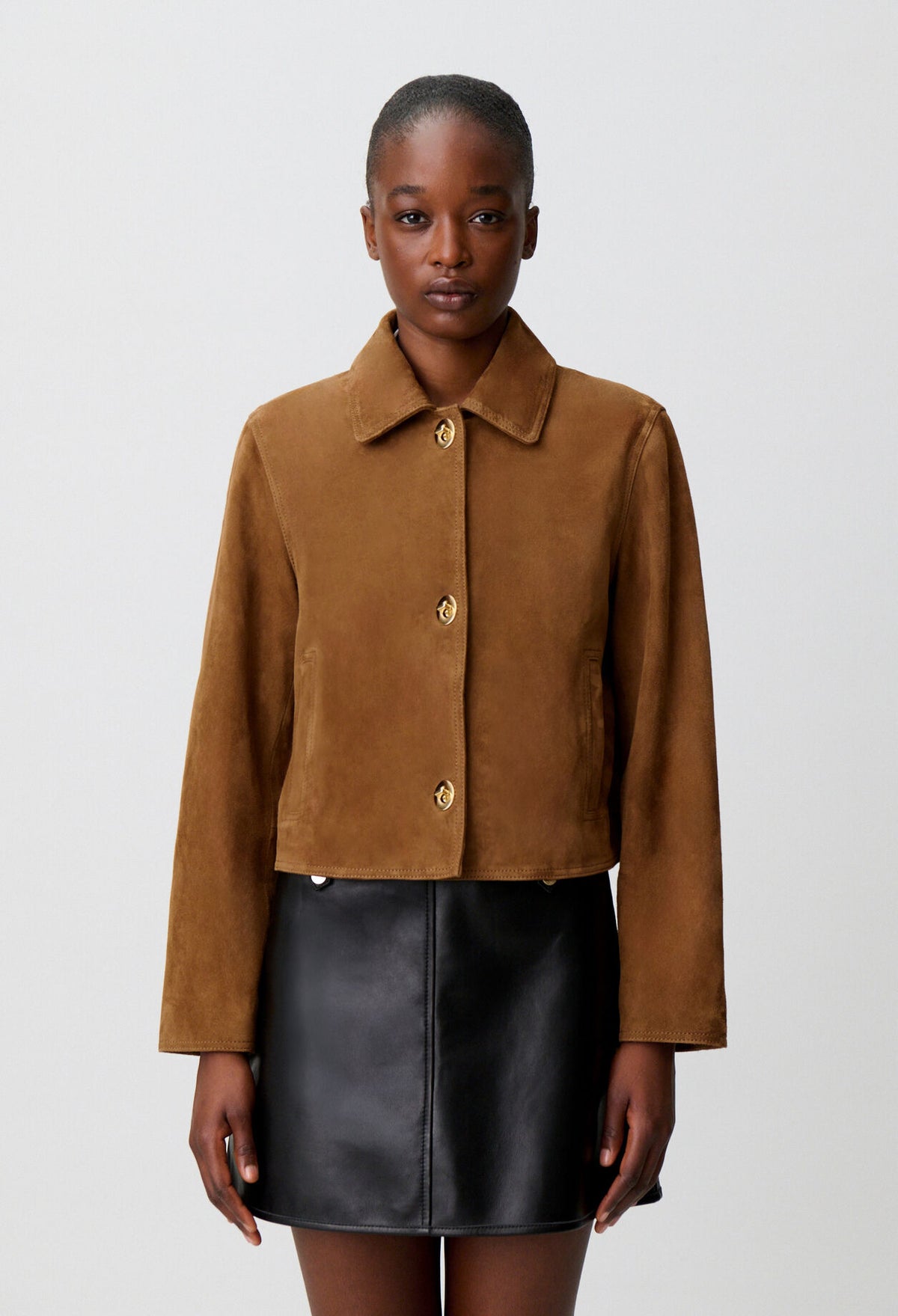 A model wearing a cropped brown suede jacket with gold buttons and a black faux-leather mini skirt, photographed against a light gray studio background.