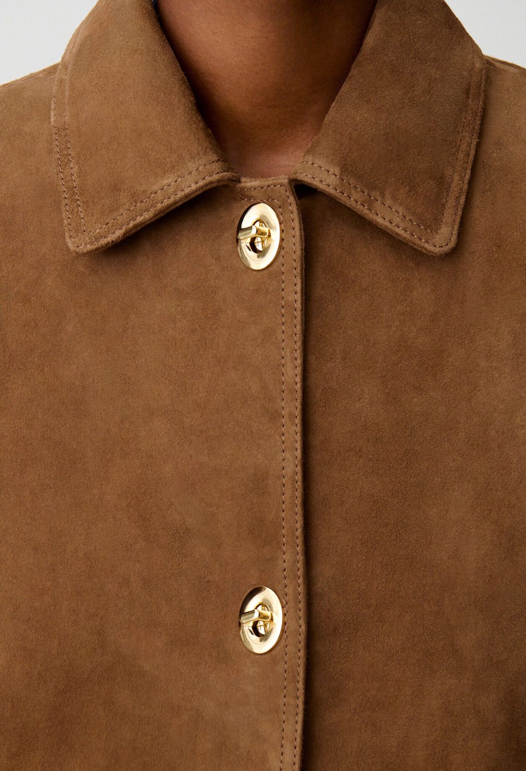 Close-up of a brown suede-like collared jacket or overshirt with gold-tone buttons, shown on a model against a light gray background.