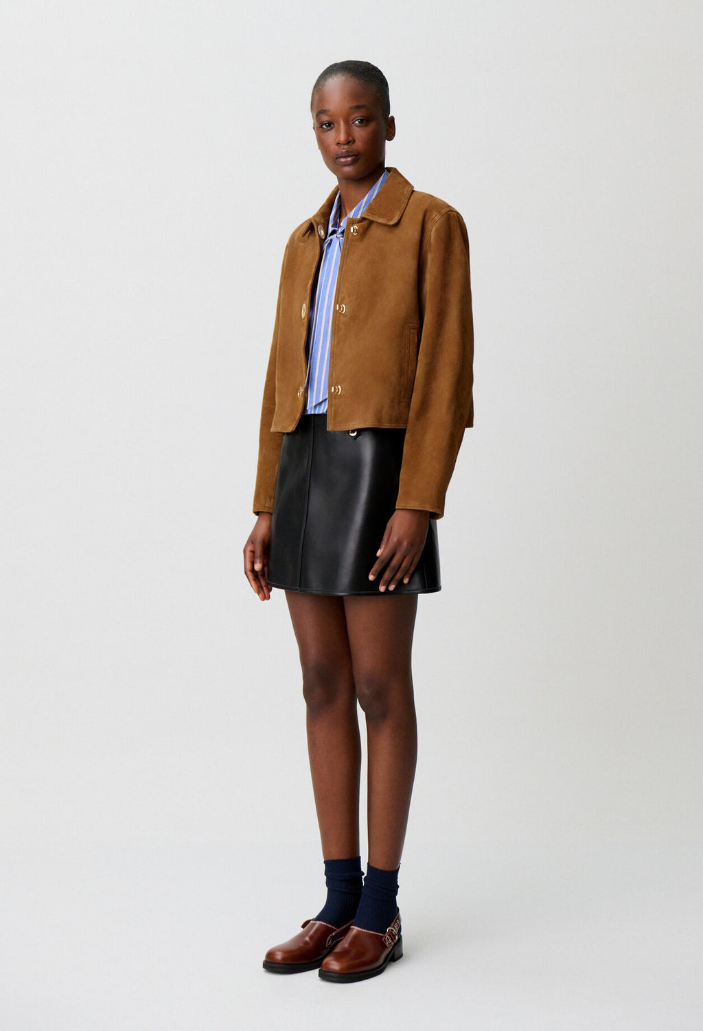 A model stands against a light gray studio background wearing a cropped brown suede jacket over a blue striped shirt, paired with a black leather mini skirt, navy socks, and brown leather loafers.